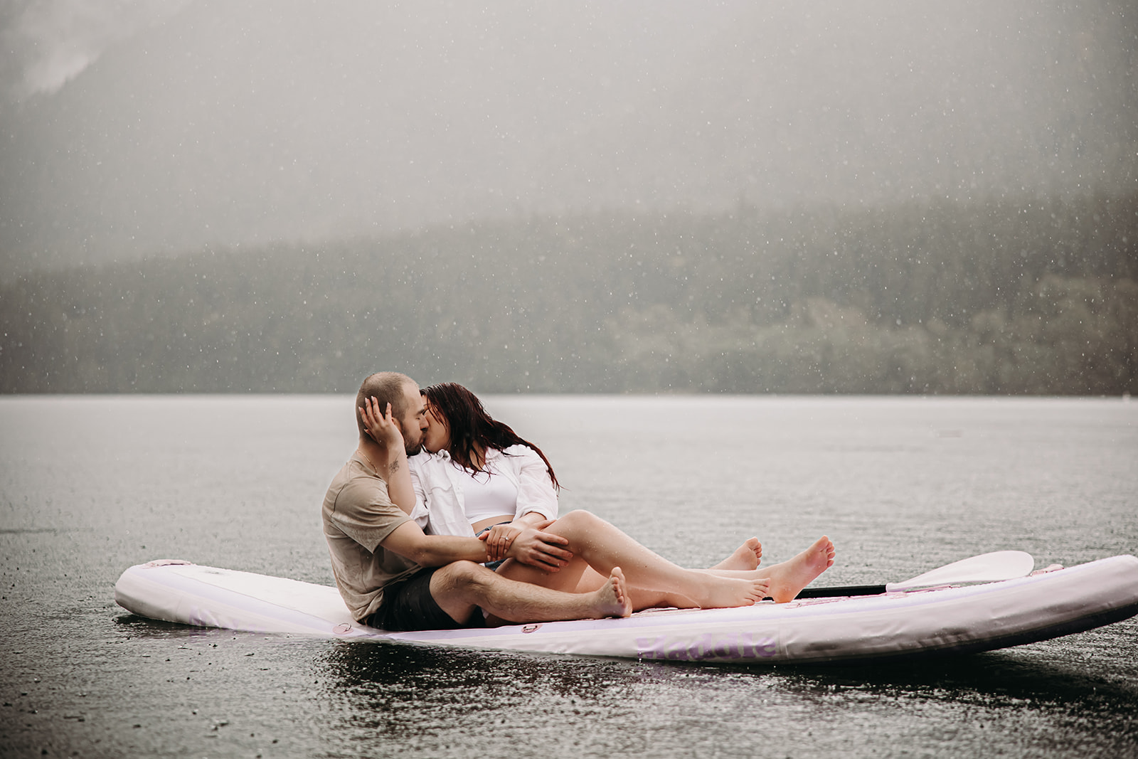 Engaged couple sharing a kiss on a paddle board during a rainy engagement photoshoot on a lake.