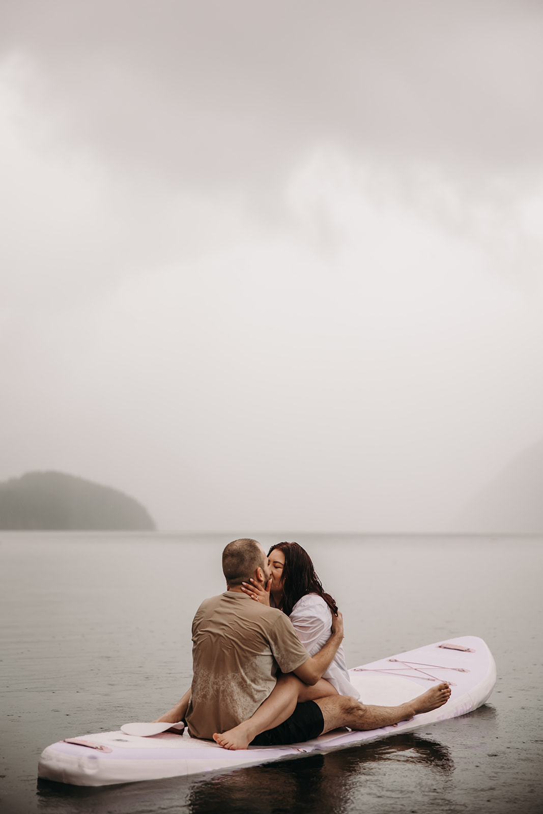 Engaged couple sharing a kiss on a paddle board during a rainy engagement photoshoot on a lake.