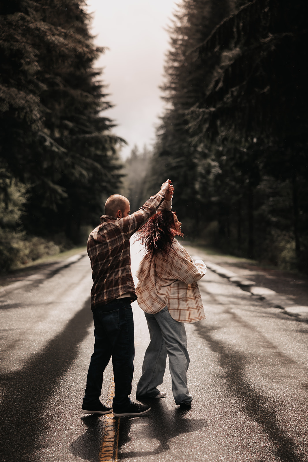 A couple twirls on a rain-slicked road during their rainy engagement photoshoot, surrounded by lush trees in the Pacific Northwest.