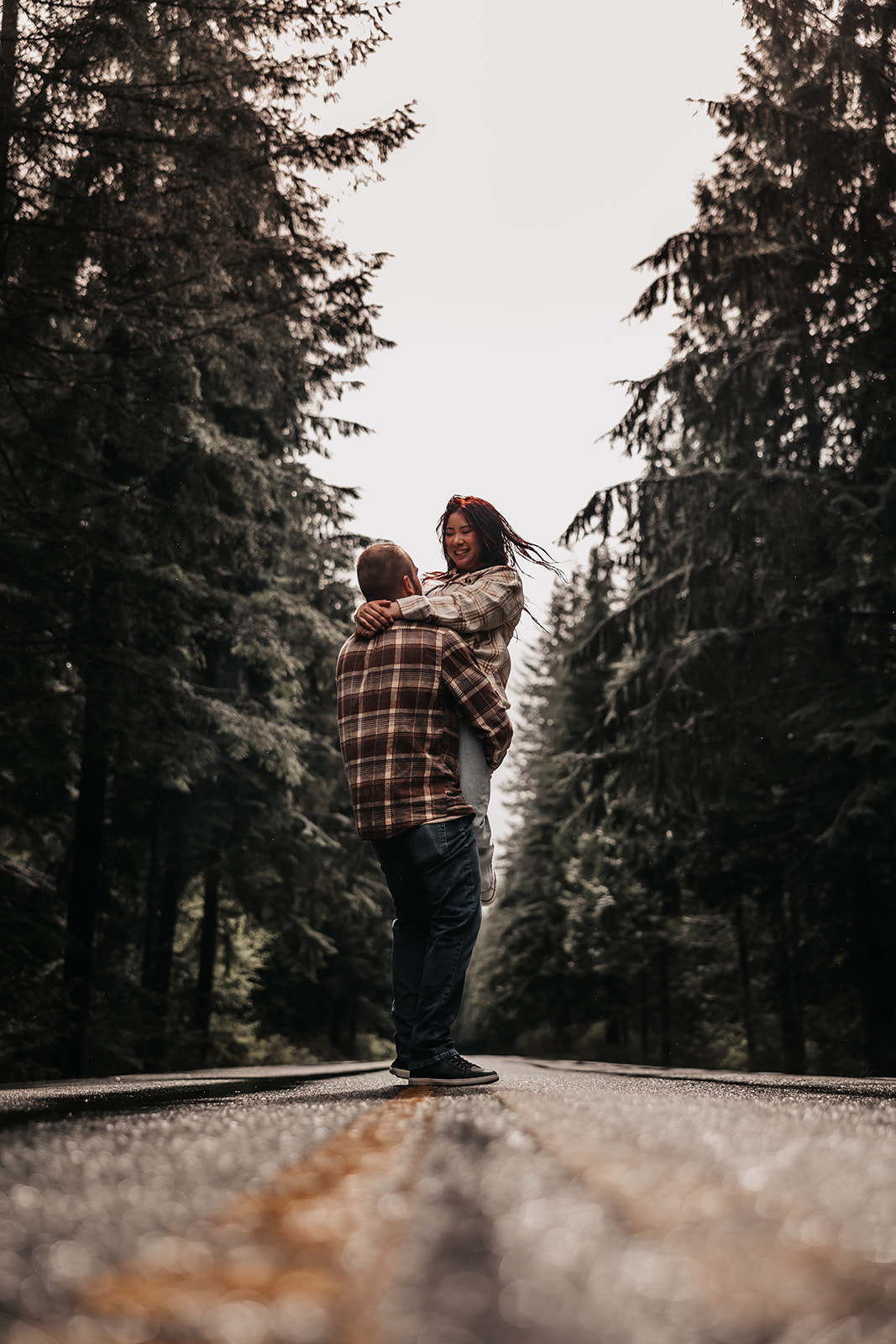 A couple shares a joyful moment on a rain-soaked road, with the woman lifted off the ground, laughing during their rainy engagement photoshoot