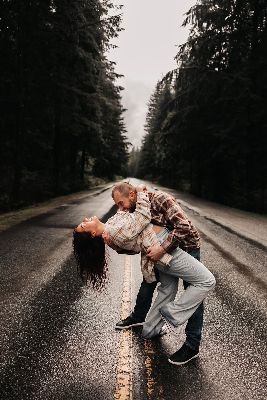 A couple twirls on a rain-slicked road during their rainy engagement photoshoot, surrounded by lush trees in the Pacific Northwest