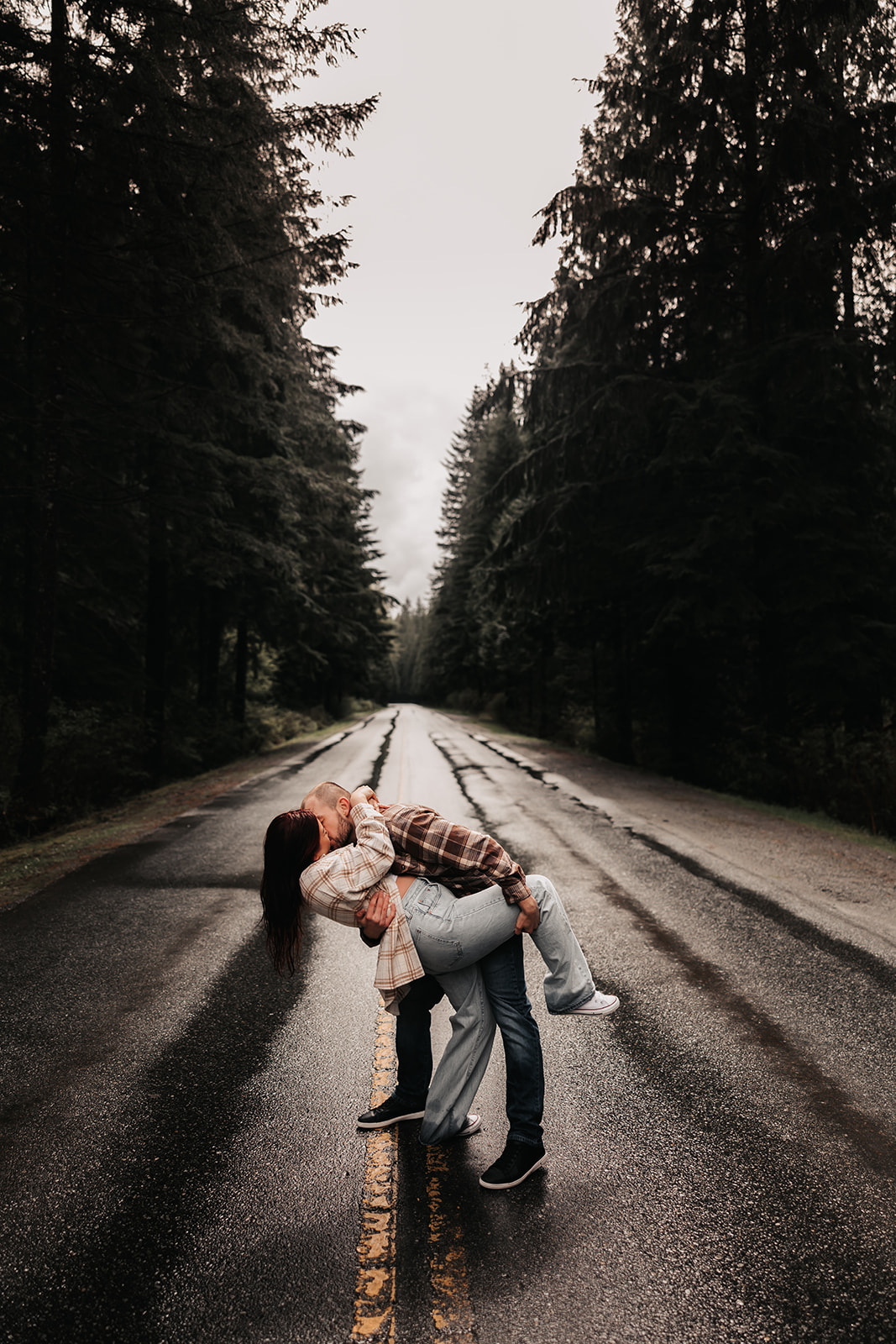 A couple passionately kisses in the rain, on a quiet road in the forest, showcasing a stunning moment in their rainy engagement photoshoot.