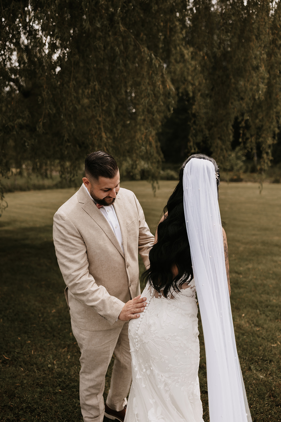 Bride looks back at her groom with a smile as he adjusts her dress, capturing the intimate moment before their ceremony begins.