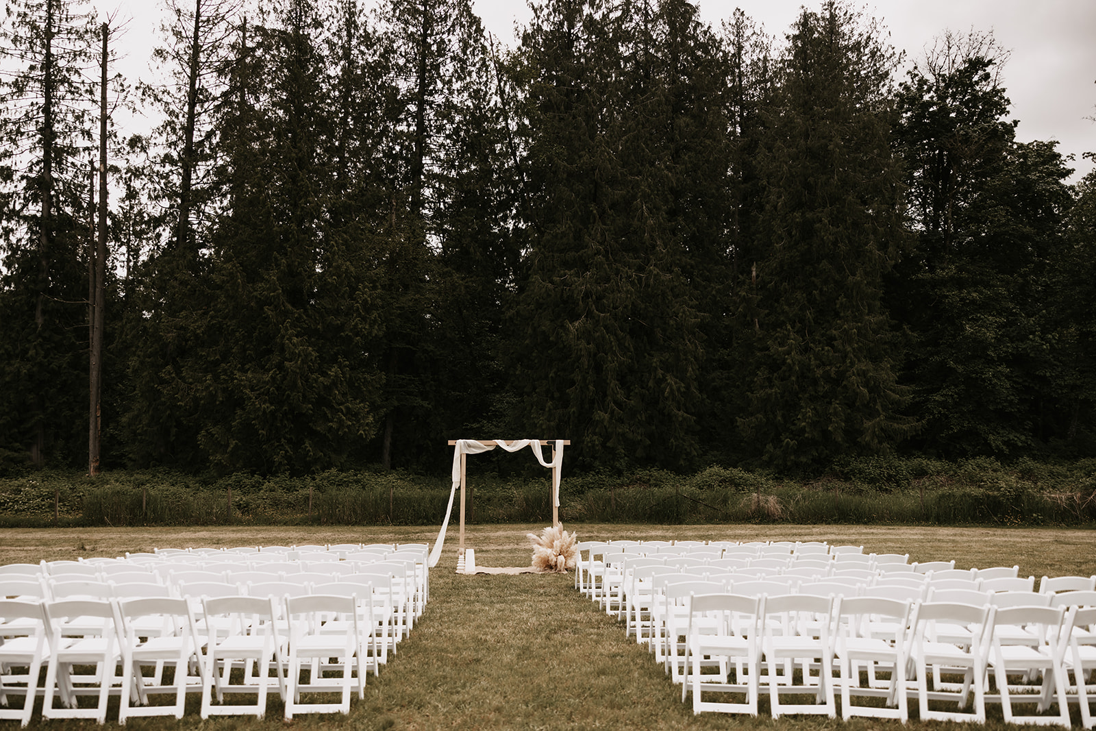 A view of the wedding ceremony space with white chairs lined up and a simple wooden arch, framed by tall trees in the background. This tranquil setting shows how to enjoy your wedding day with a peaceful, natural backdrop.