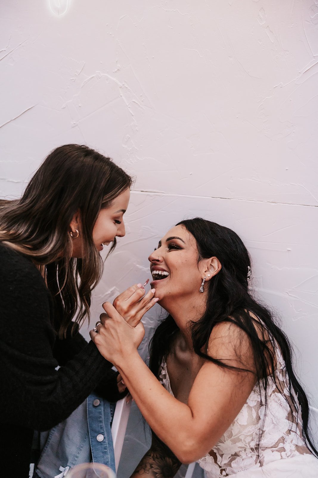 A bride and her close friend laughing while the friend helps her with her lipstick touch-up, capturing a candid and intimate moment between the two. A reminder of the small, meaningful moments that make your wedding day special.