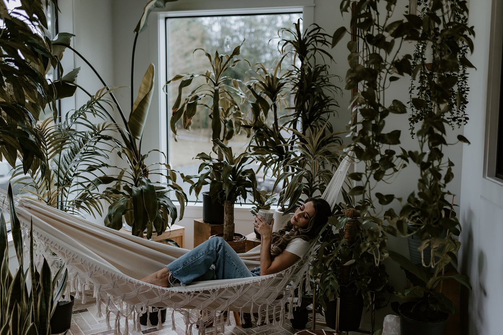 A woman lounging in a hammock surrounded by indoor plants, sipping a cup of tea or coffee, offering a cozy vibe perfect for fun engagement photo ideas.