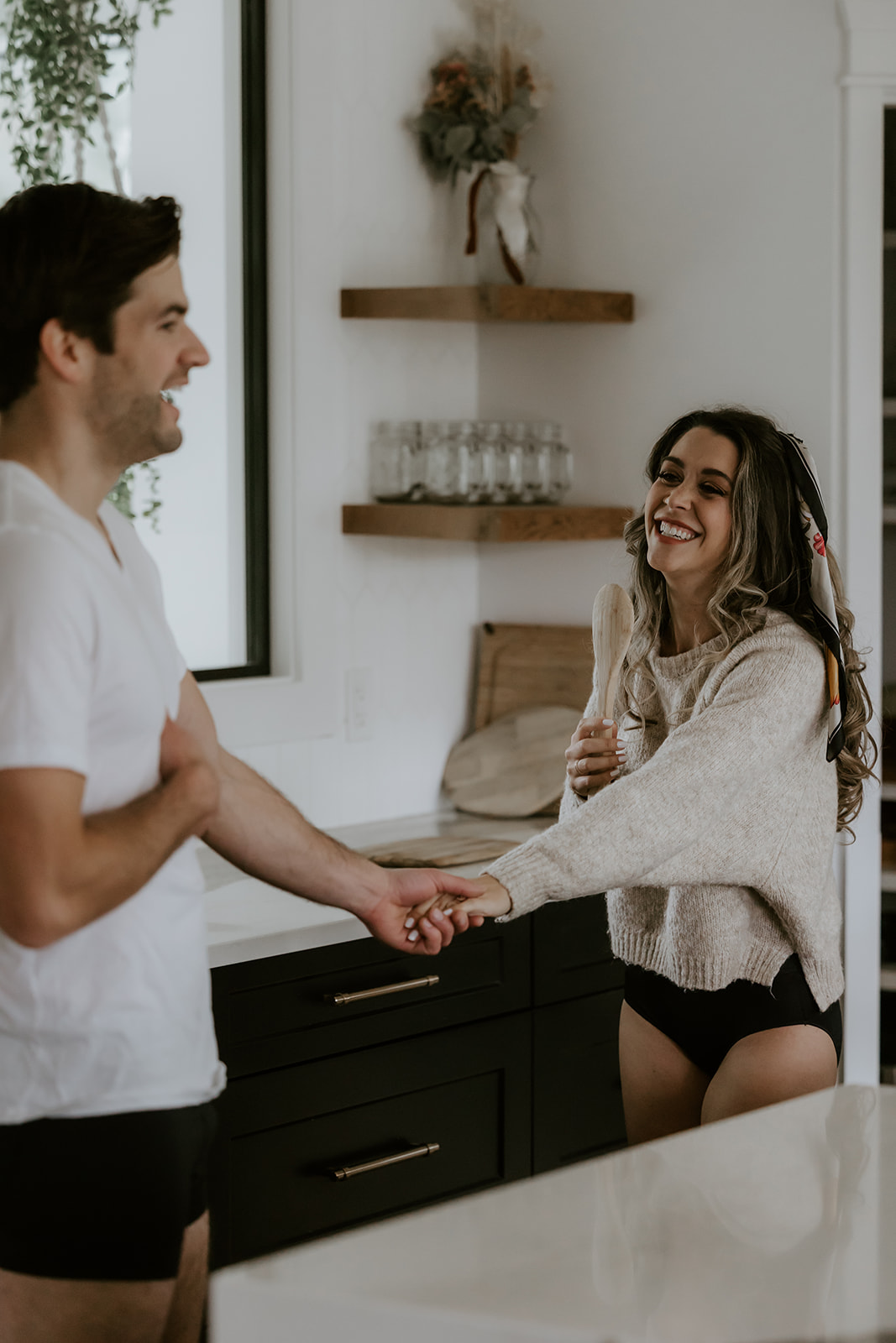 A couple laughing and sharing a sweet moment in the kitchen while holding hands and preparing to cook together in casual clothes, creating candid, fun moments.