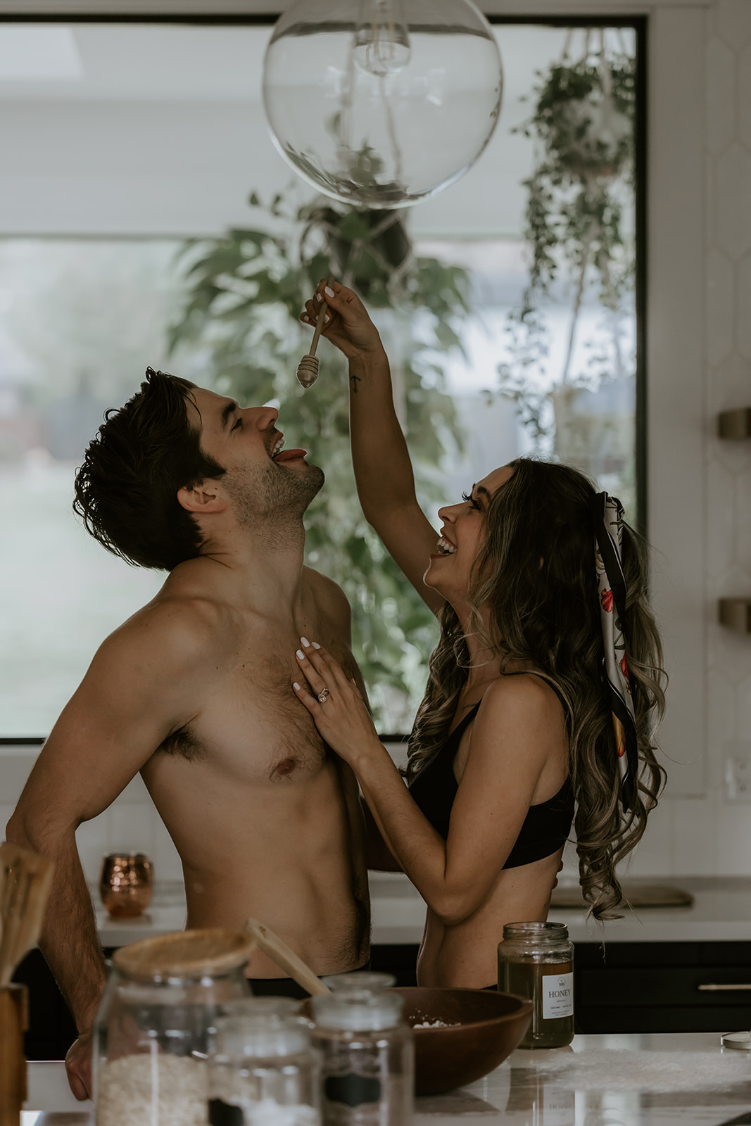 A couple feeding each other honey in the kitchen while laughing, creating a sweet and intimate moment during their engagement shoot at home.