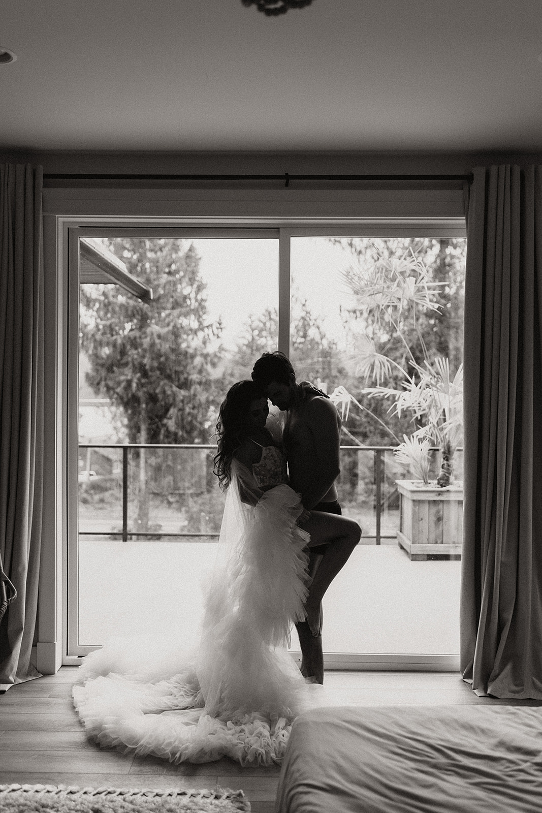 A couple shares a joyful moment while standing in front of a large window with dramatic lighting, creating an intimate vibe for their fun engagement photo ideas.