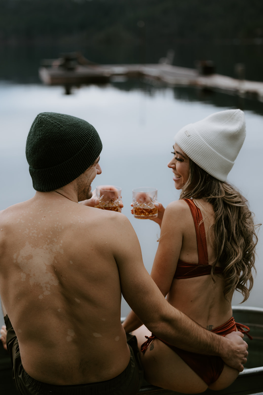 A couple enjoying the outdoors, sitting together on a dock by a serene lake, with glasses of whiskey in hand. This cozy moment captures their relaxed vibe with fun engagement photo ideas, set against the tranquil water and mountains in the background.