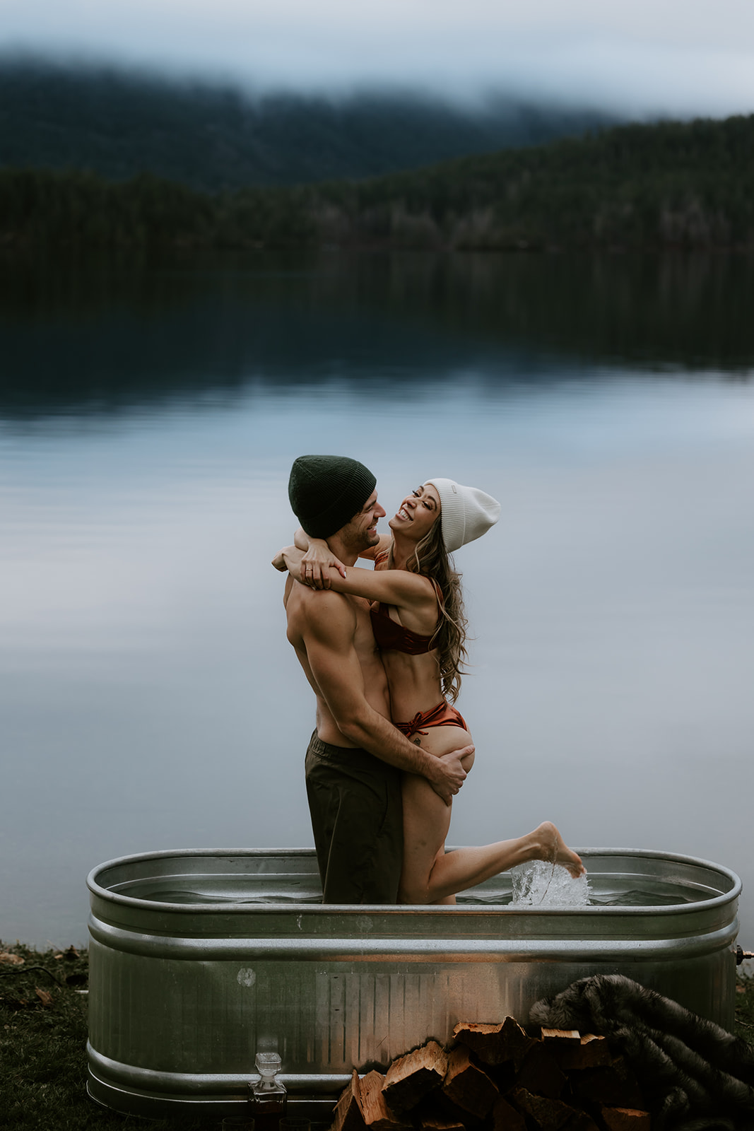 A playful and intimate shot of the couple as one lifts the other out of a rustic metal tub by the lake. The couple's matching winter hats and warm smiles create the perfect atmosphere for fun engagement photo ideas that embrace adventure and love in the outdoors.