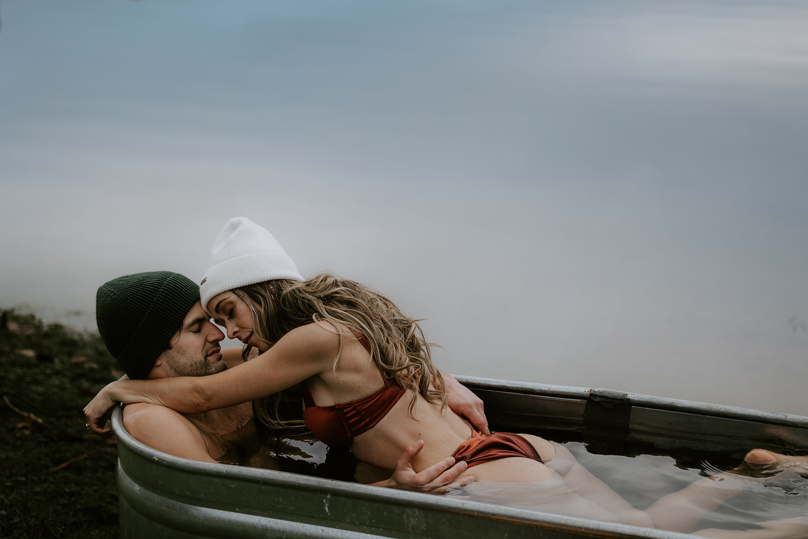 The couple shares a loving moment while relaxing in a metal tub beside a lake, their faces close as they gaze at each other. The cool, misty atmosphere makes this image a beautiful example of fun engagement photo ideas that capture raw affection and adventure.