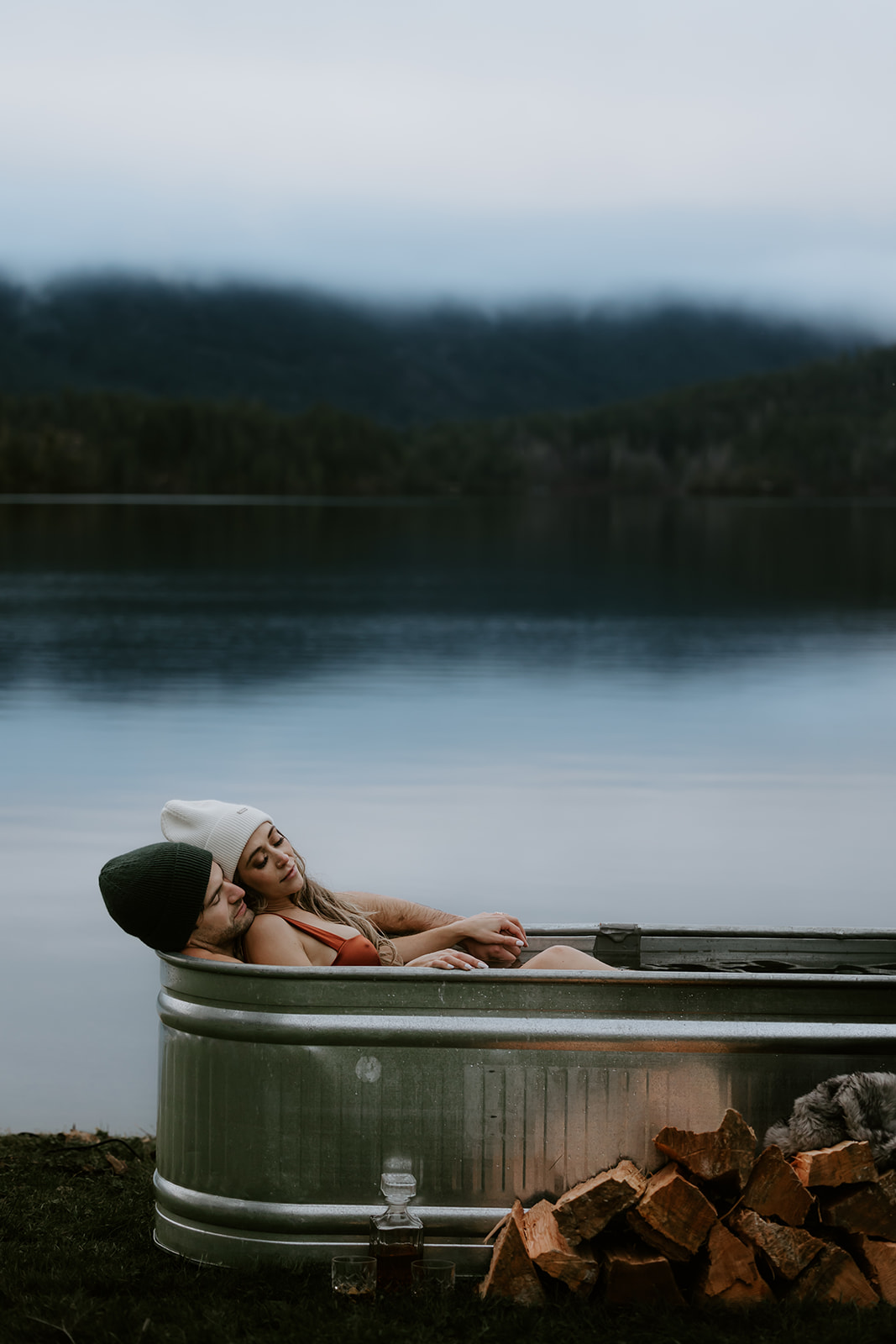 In a cute and candid pose, the couple shares a sweet moment, sitting together in a water-filled metal tub. Their cozy winter hats and the serene lake backdrop are perfect for fun engagement photo ideas that highlight their deep connection.