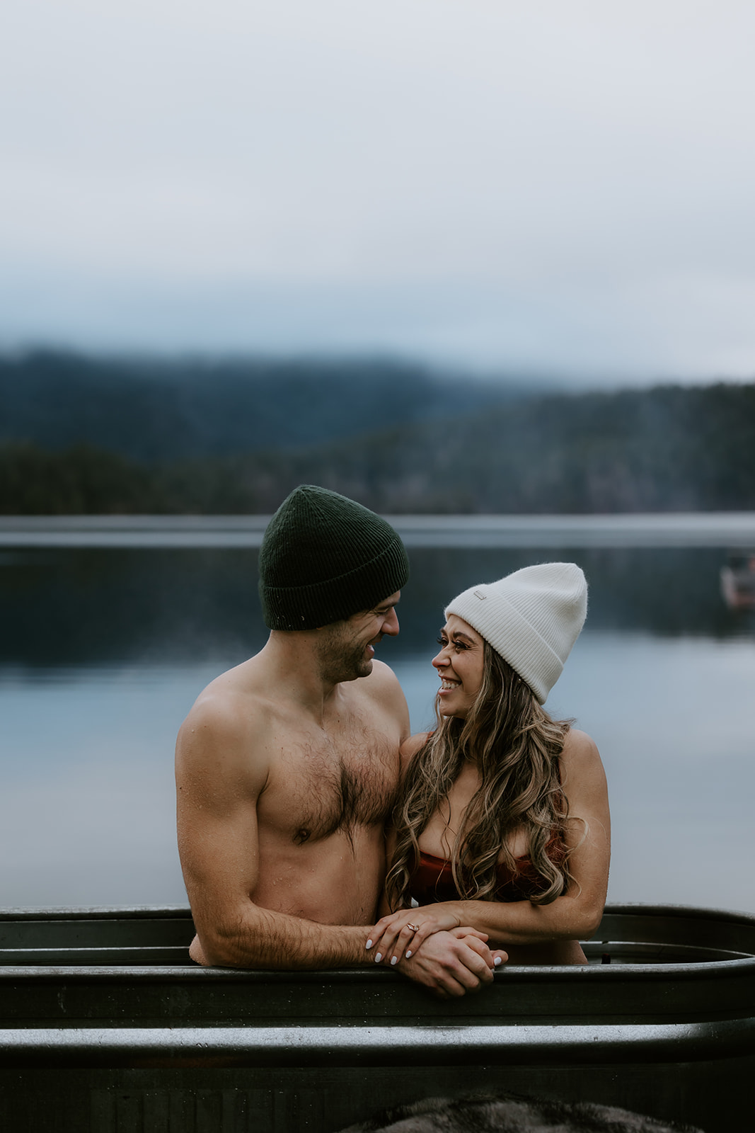 The couple shares a playful moment in the metal tub, laughing and looking into each other's eyes. The lake view and serene environment make it an ideal setting for fun engagement photo ideas that capture carefree love and a sense of adventure.