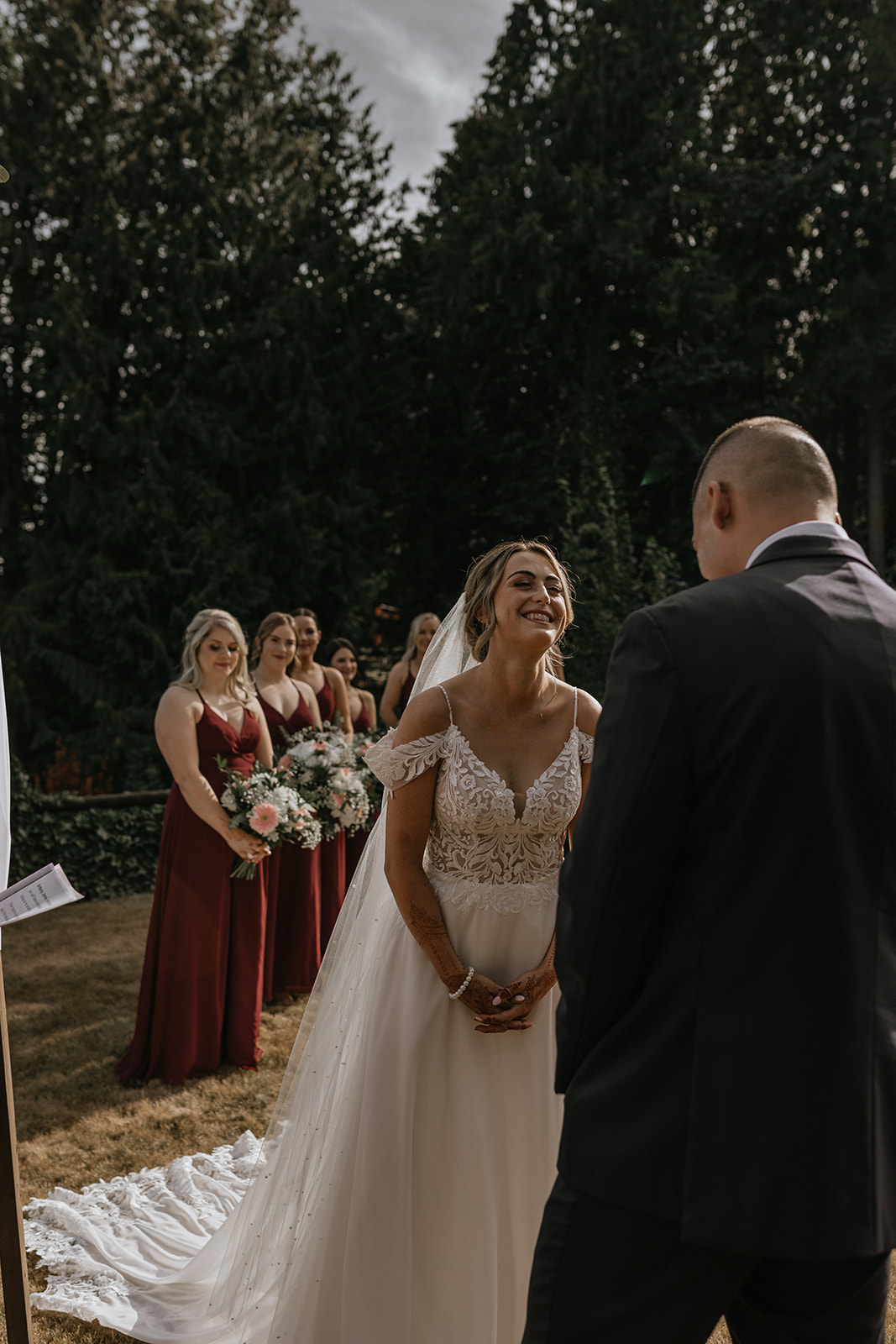 The bride smiles at her groom during their backyard wedding ceremony, with the bridal party looking on in the serene Fraser Valley.