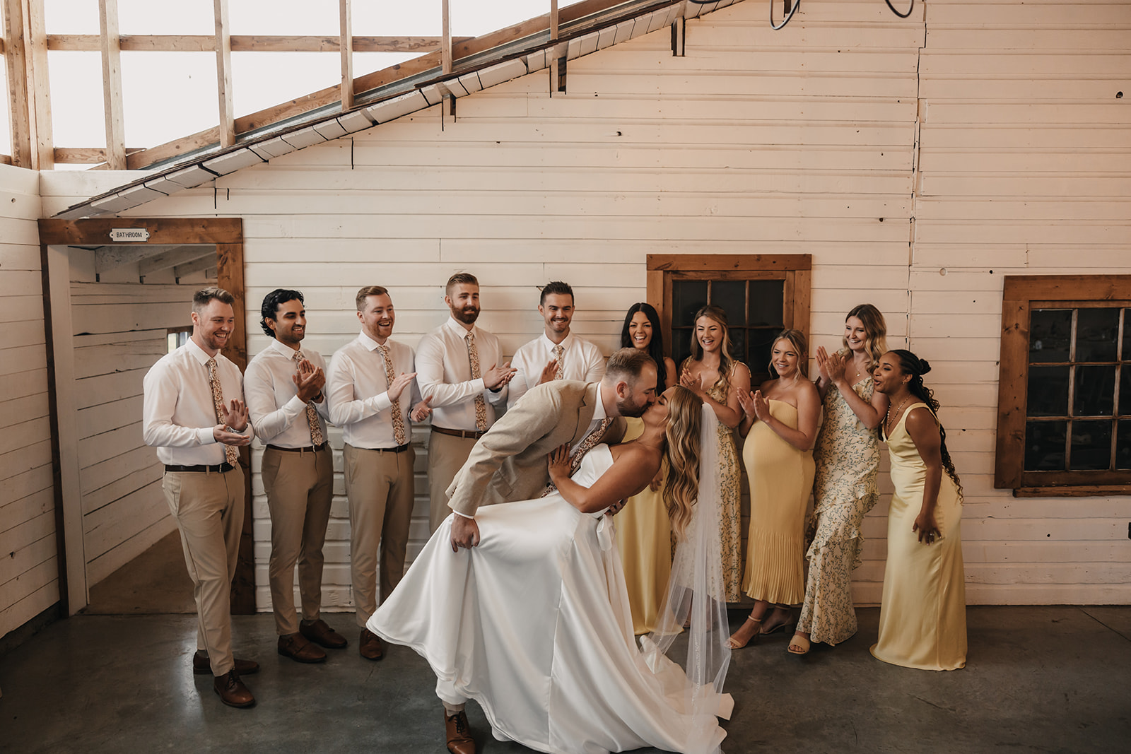 A joyful moment with the couple kissing while their bridal party watches and cheers them on in the background, all captured inside a rustic barn setting.