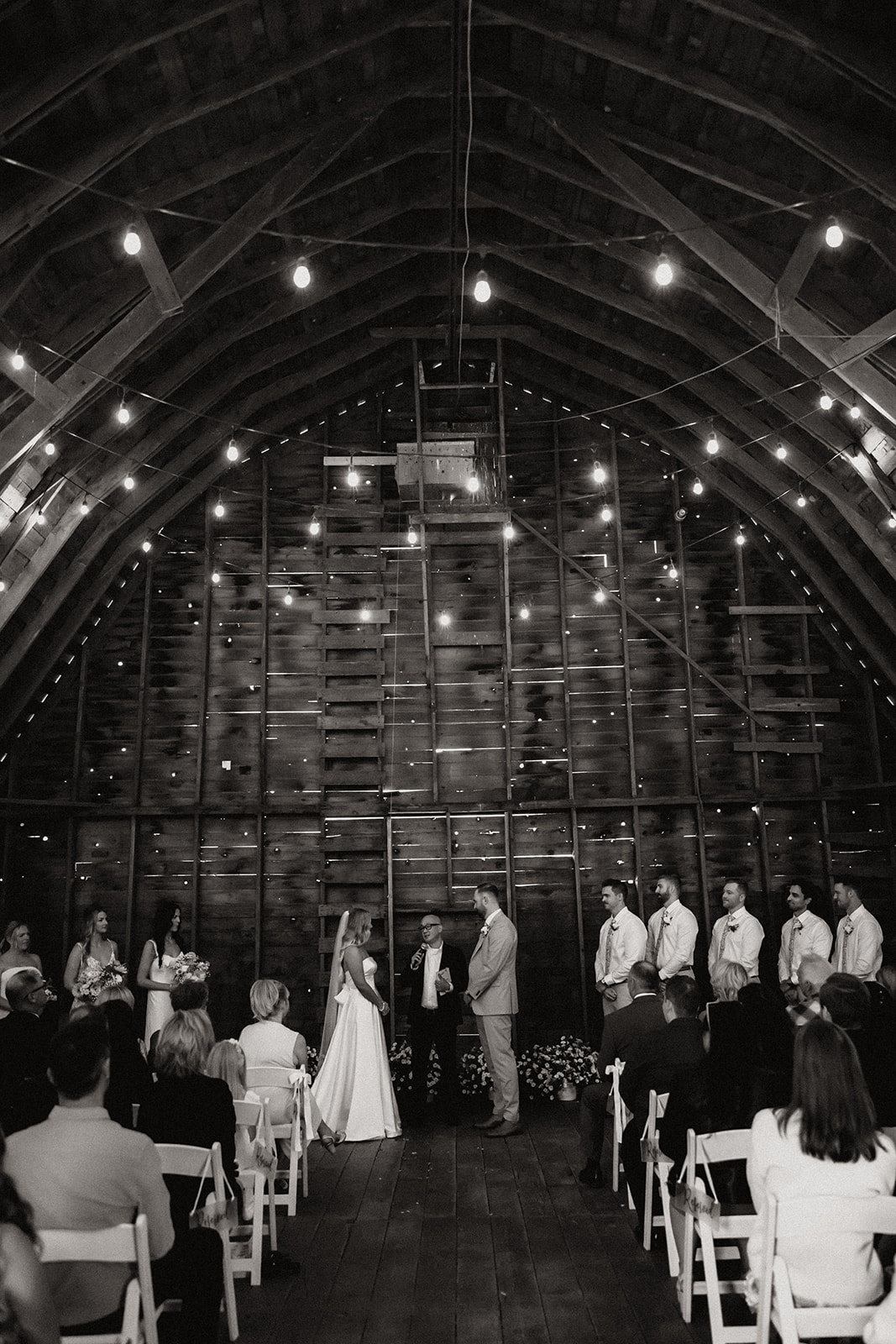 A wide view of the ceremony in a barn with glowing string lights. The bride and groom’s ceremony showcases a stress-free wedding day, surrounded by love and laughter.