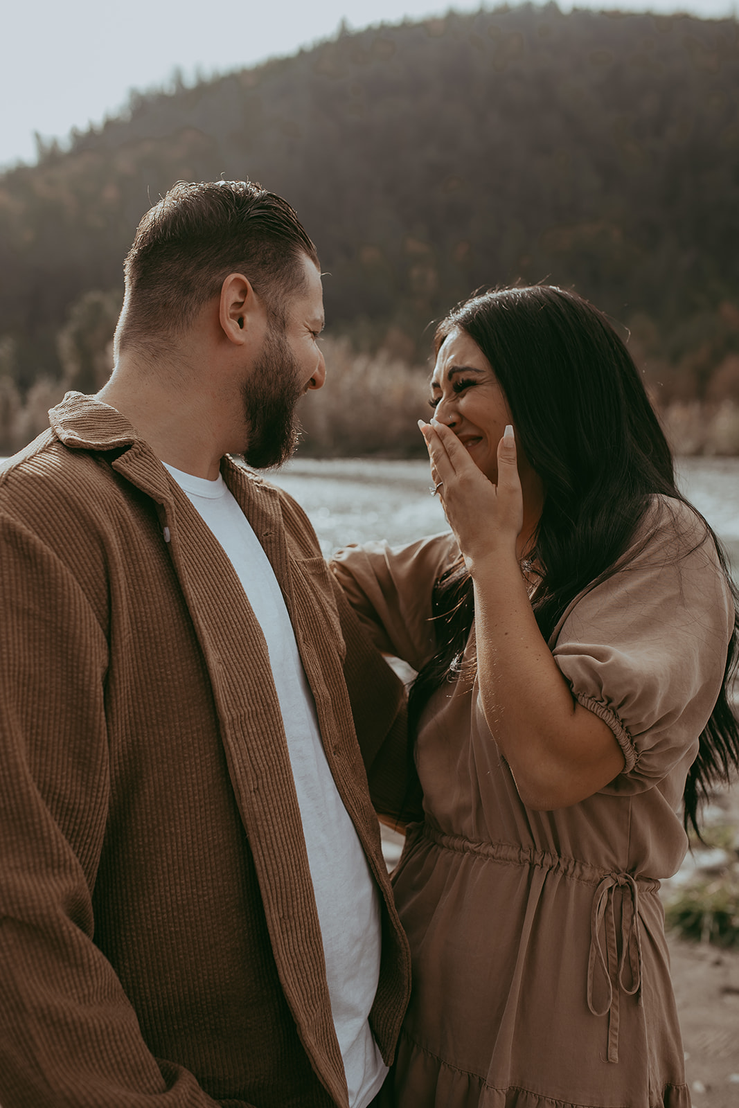 Couple laughing and having fun together during their engagement session by the water, enjoying a natural moment.