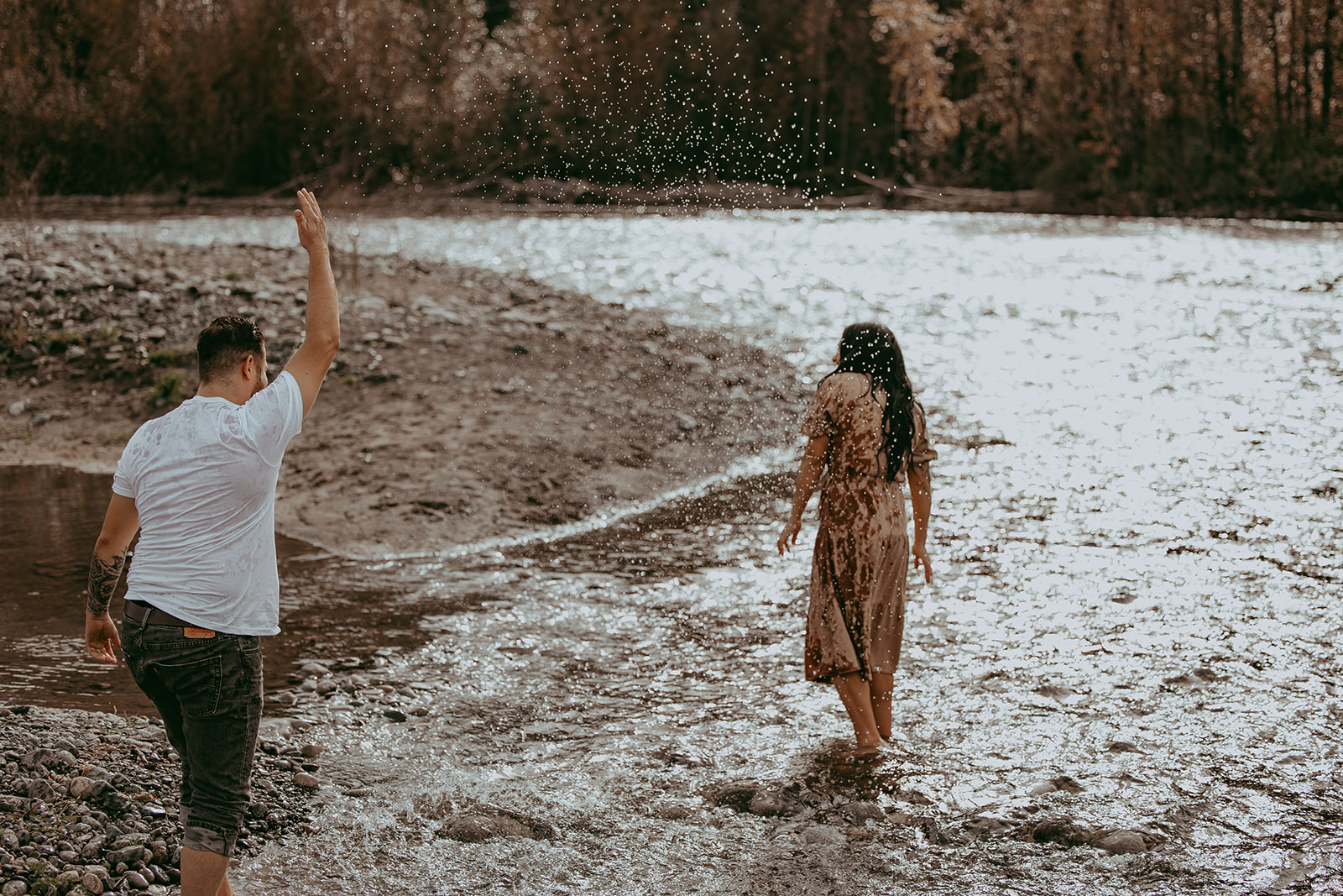 A couple enjoying their engagement session by the river, with the man playfully tossing water into the air while the woman walks ahead, smiling.