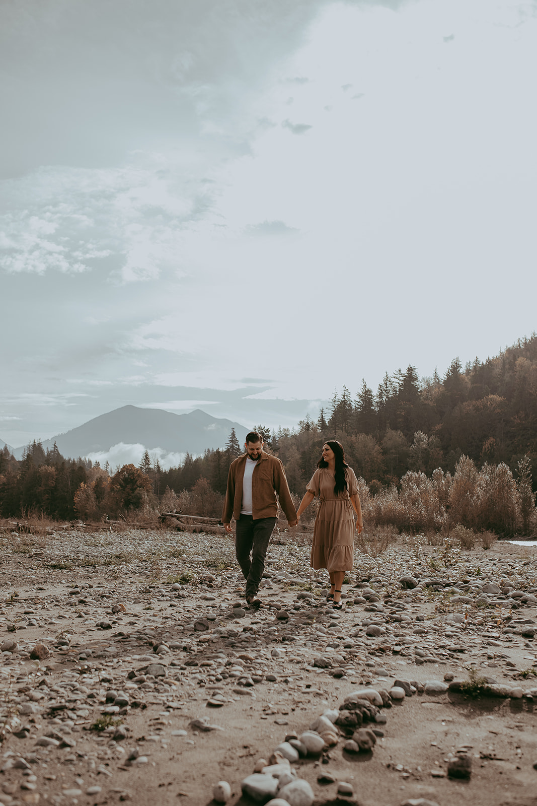 The couple walking hand in hand along a riverbank, with scenic mountains and trees in the background, capturing the essence of their engagement session.