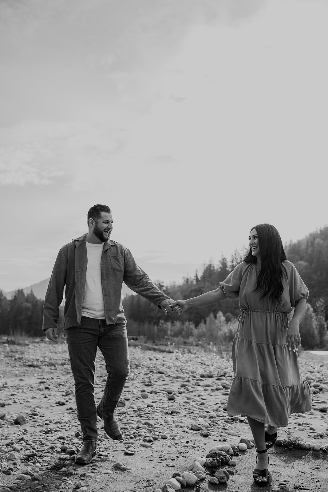 A black-and-white image of the couple walking hand-in-hand, smiling and laughing during their engagement session by the river.