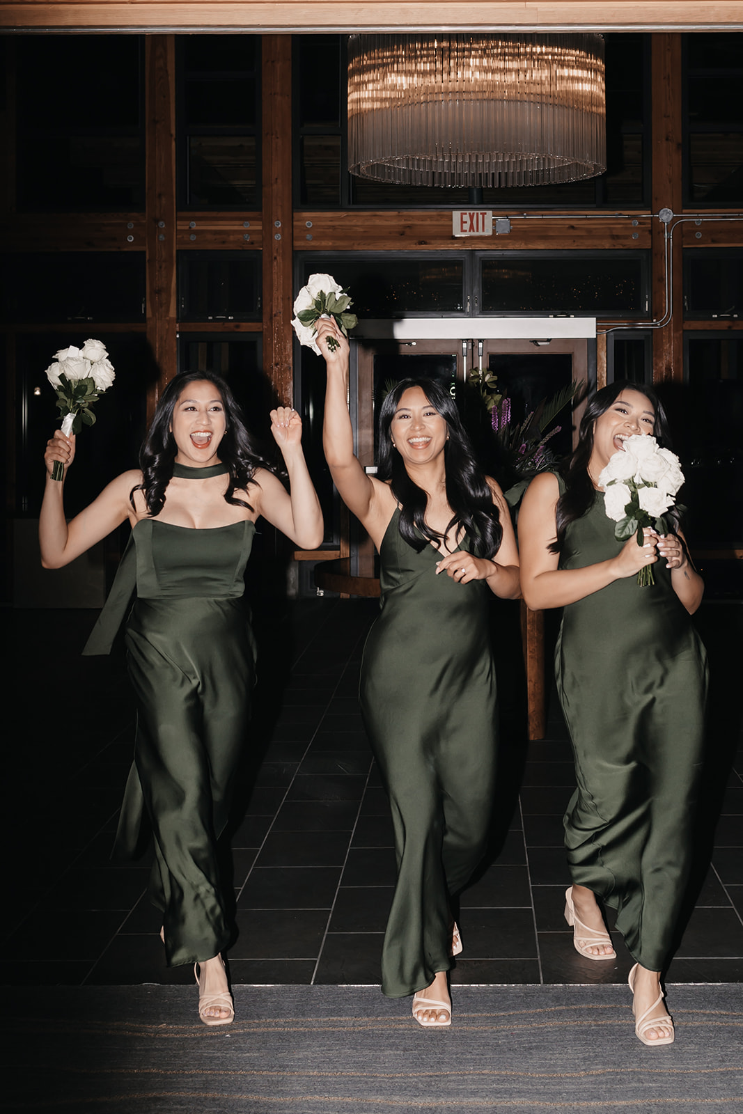 Bridesmaids joyfully celebrating with white rose bouquets, walking down a hallway at the reception venue. This moment would make a fun and lively addition to your wedding day timeline.