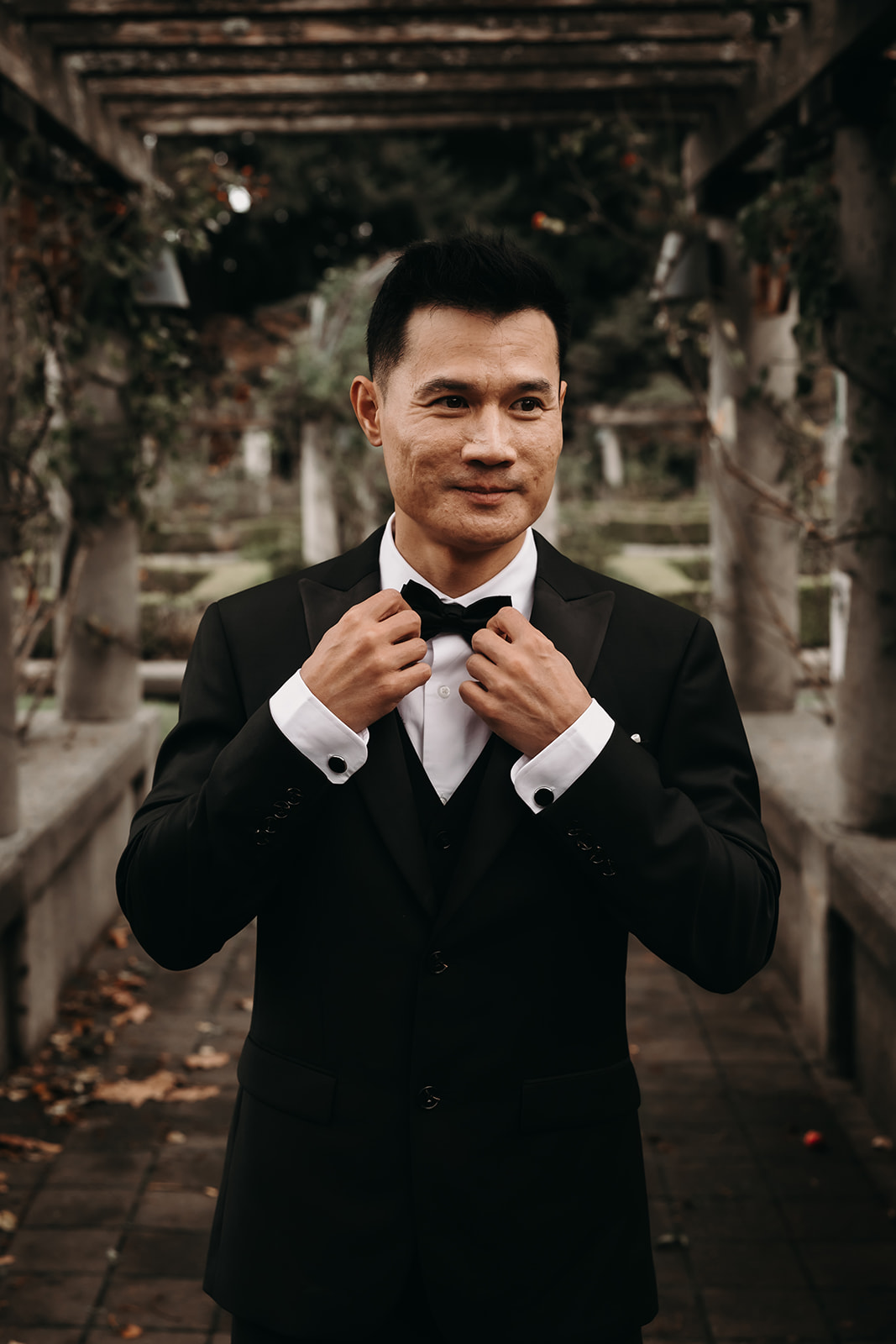 Wedding Day Timeline - Groom standing alone in a garden pathway at the venue, adjusting his bow tie before the ceremony starts, looking sharp and ready for the big day.