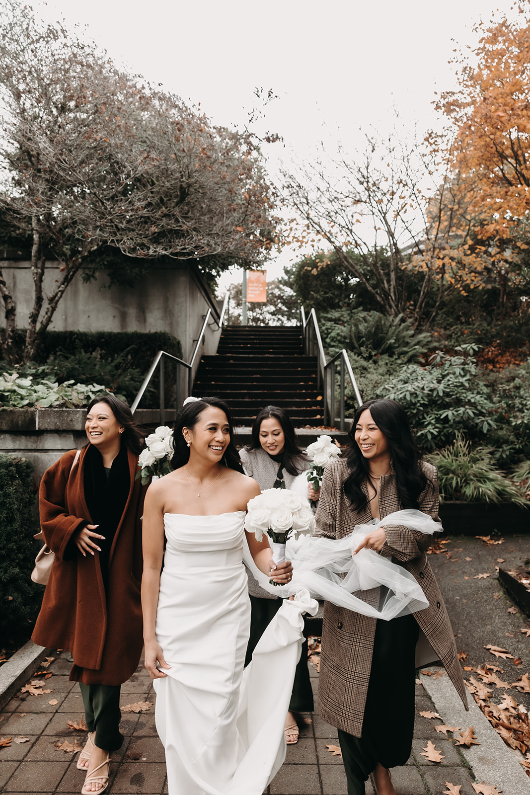 Bride and bridesmaids walking to the ceremony venue, smiling and carrying bouquets of white roses, with fall foliage in the background at a Vancouver wedding.