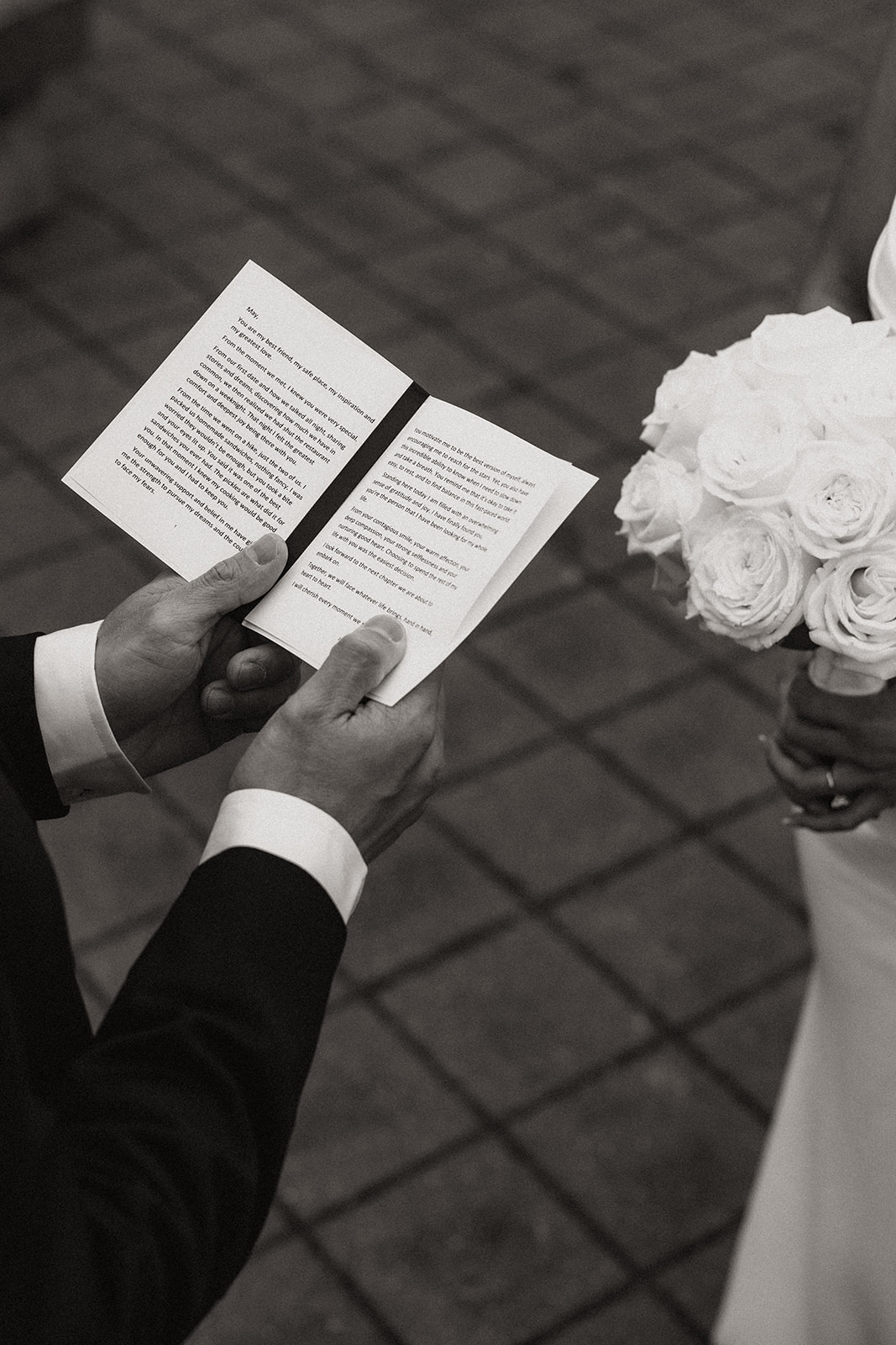 Close-up shot of the groom reading from a book during the ceremony. His hands hold the pages as the bride stands in front of him, her white bouquet held gently. The soft focus on the book emphasizes the meaningful words spoken during this intimate part of the wedding day timeline.