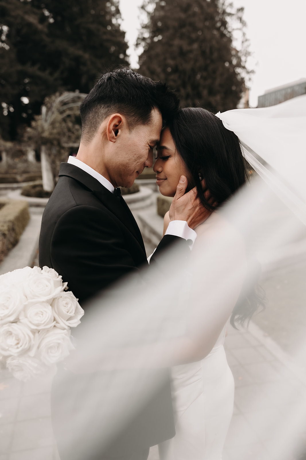 Bride and groom embrace with the bride’s veil flowing behind them. As the veil moves in the breeze, this moment captures the joy and spontaneity that can be planned into a wedding day timeline. Their love shines through as they embrace their first kiss as a married couple.