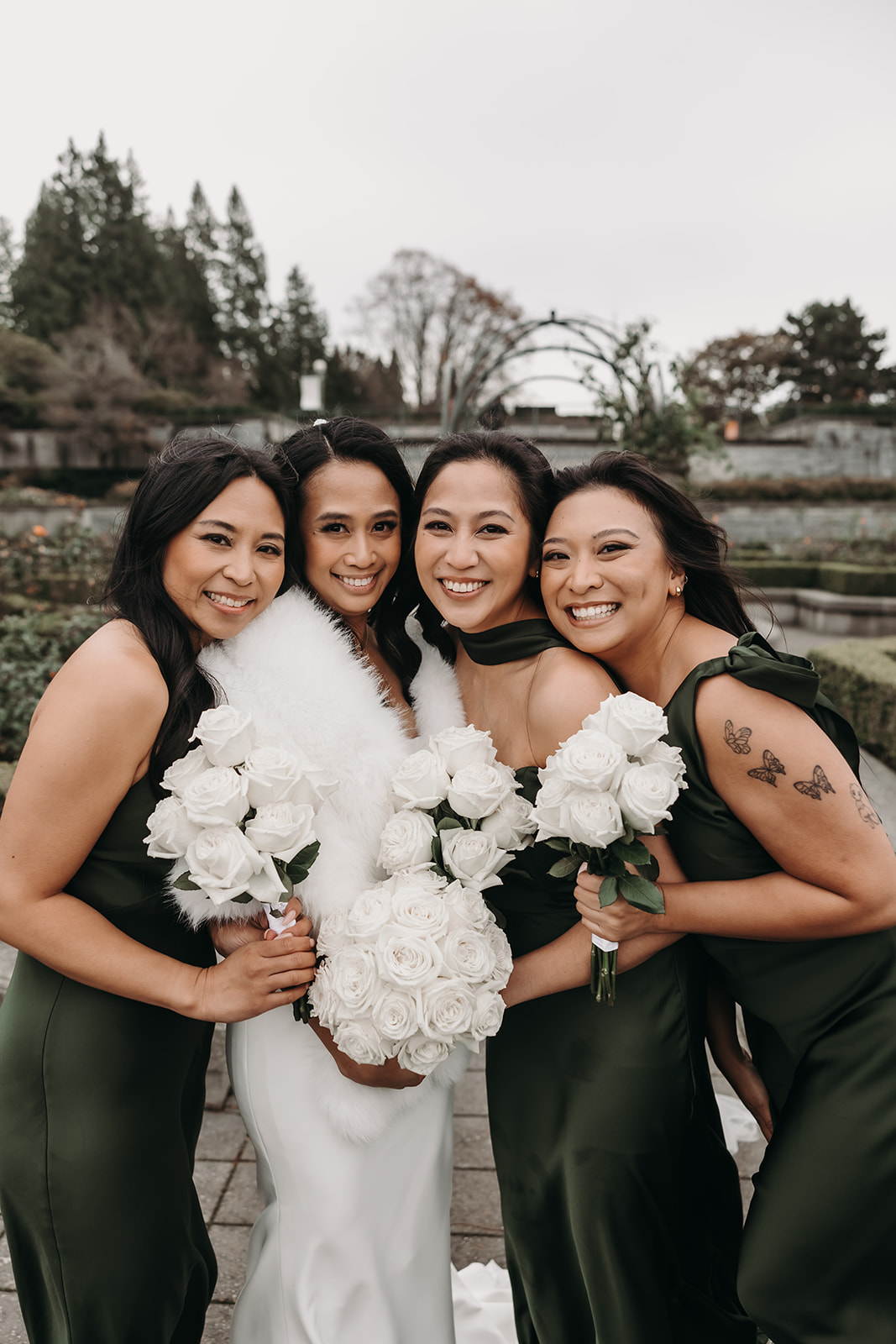 Bride and her bridesmaids laughing and posing for a photo. The bridesmaids, all wearing deep green dresses, hold bouquets of white roses. The image captures the essence of friendship and joy, showcasing how the wedding day timeline incorporates moments of laughter and connection.