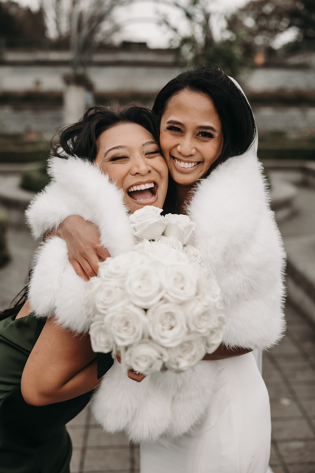 Bride and bridesmaid share a joyful embrace, laughing together. The bride and bridesmaid, both wearing luxurious white fur wraps, stand in front of the garden, their smiles wide and genuine. This moment is another example of how a well-planned wedding day timeline can capture the emotional support shared between loved ones.