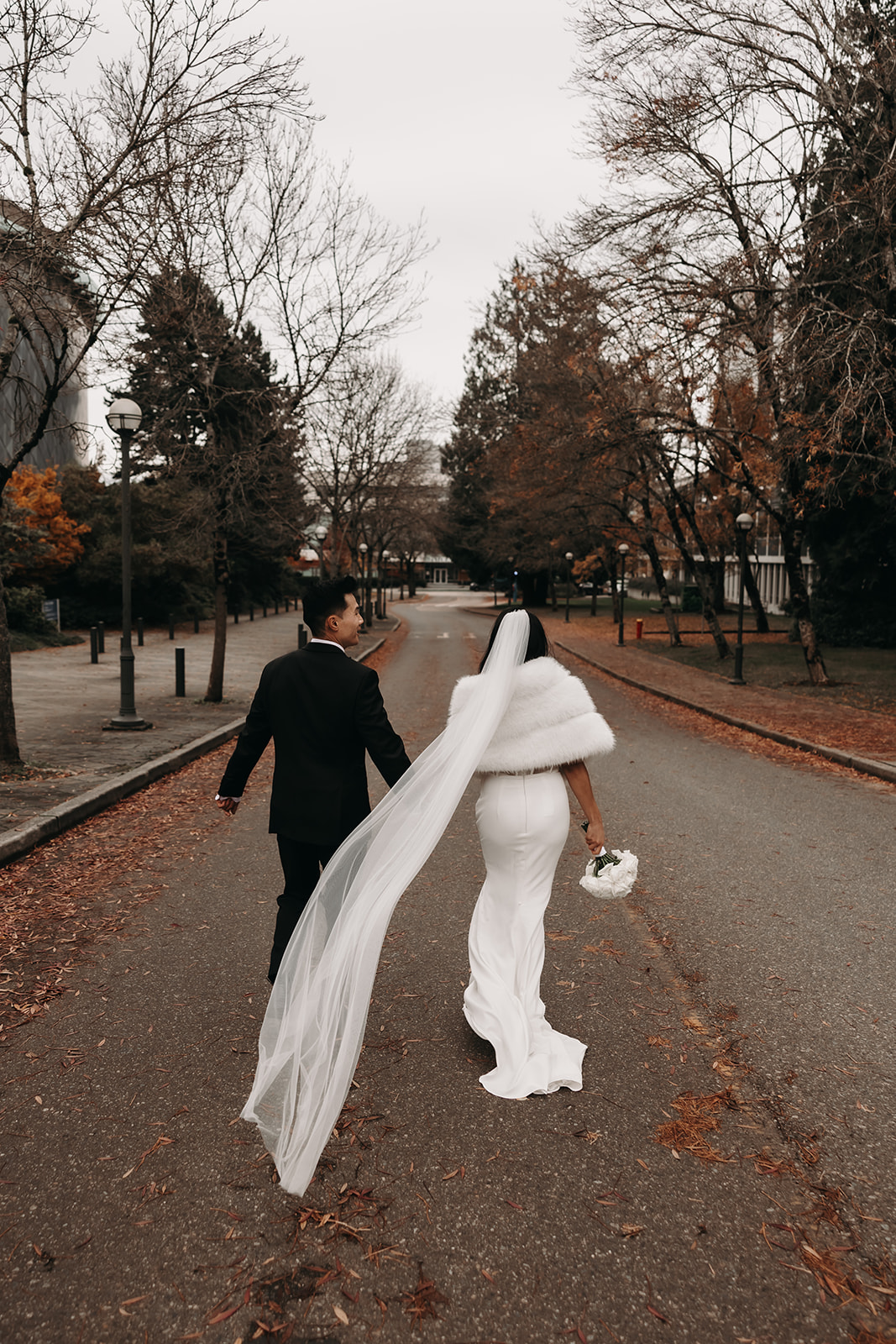 Couple holding hands while walking down a path surrounded by trees and holding a bouquet of white roses, with the bride's veil flowing behind.