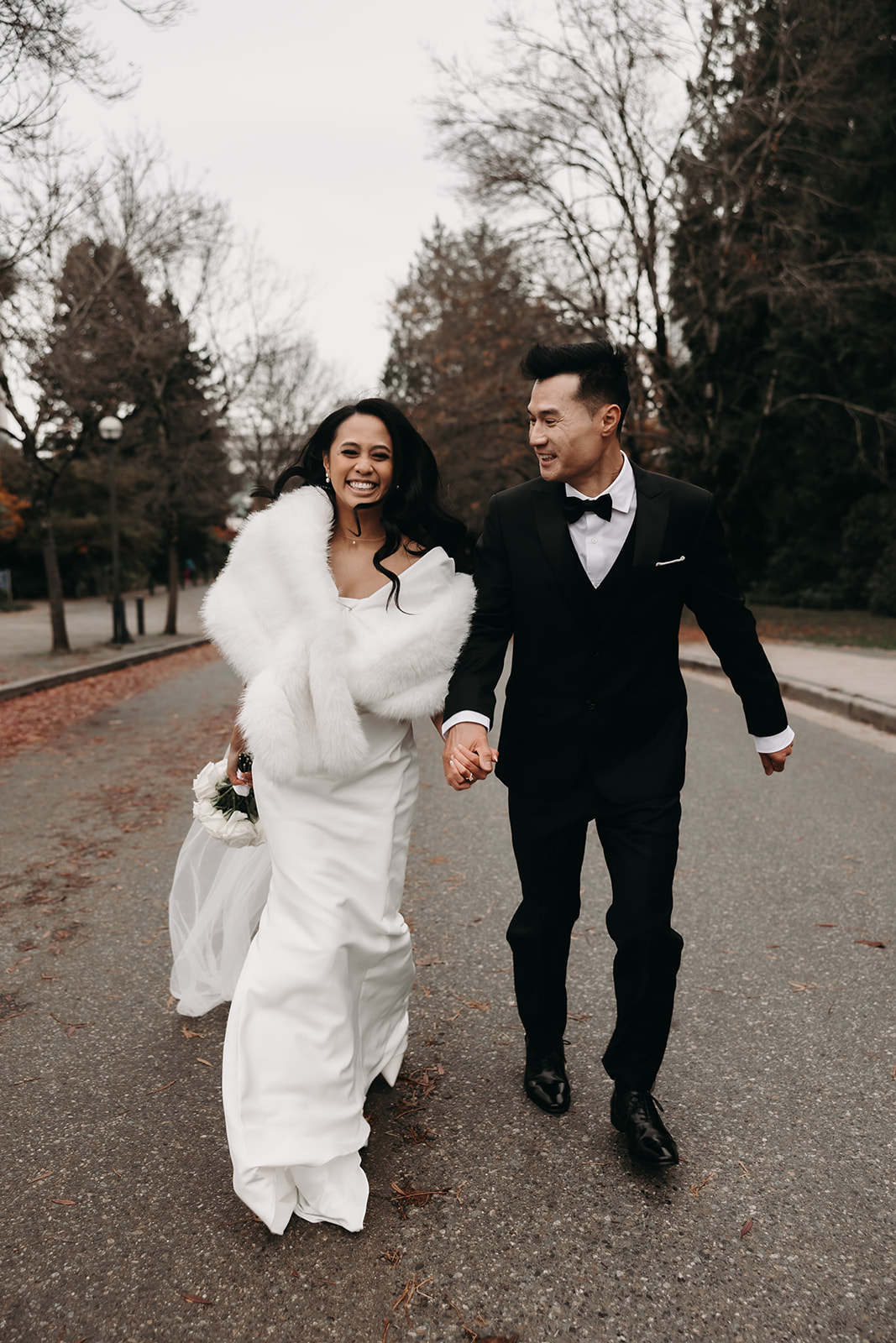 Bride and groom walking down the path laughing and holding hands, the bride's white fur stole adding a touch of elegance to her bridal gown.