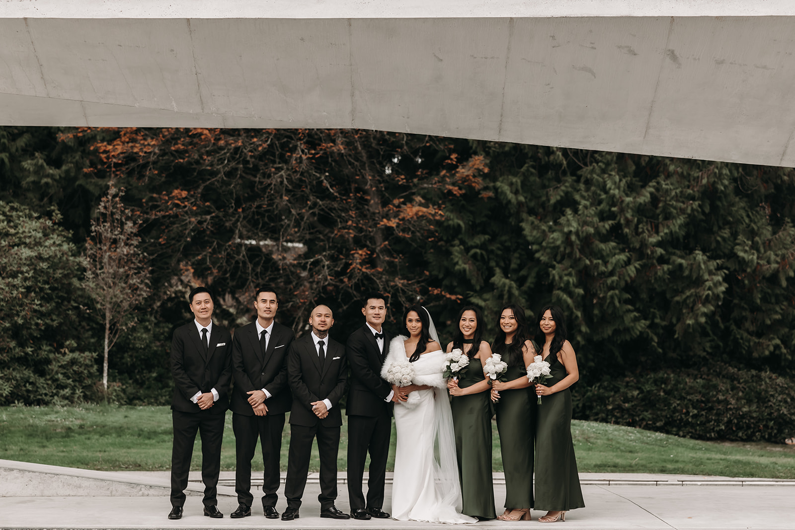 Bridal party posing together under a modern architectural structure with the groom and bride in the center, both holding bouquets of white roses.