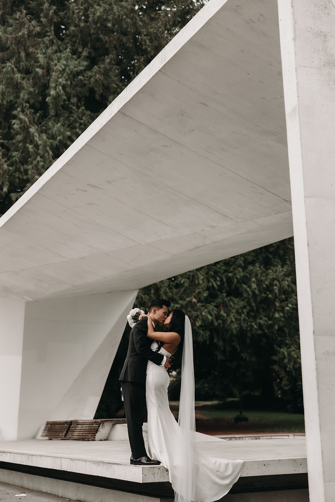 Bride and groom sharing a kiss underneath an architectural structure, with the bride’s veil floating in the air as they embrace.