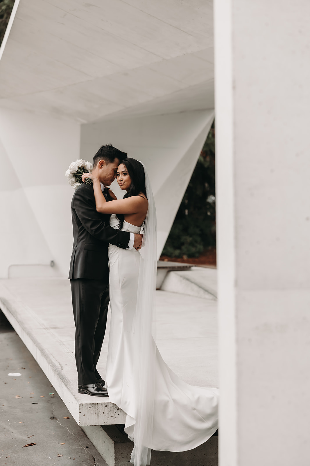 Bride and groom embracing under the sleek structure, their faces close together, while the bride's veil adds a romantic touch to the image.