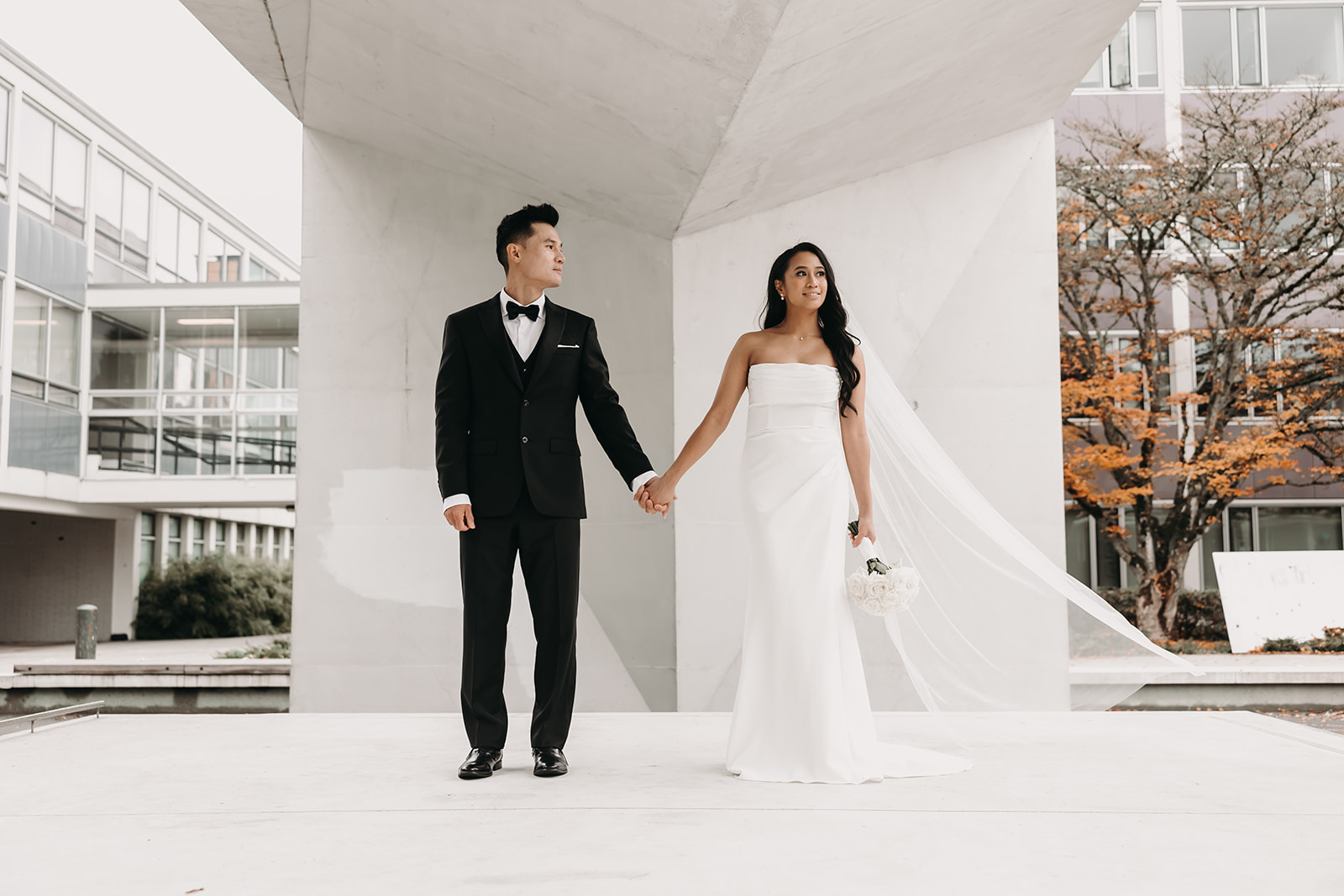 Bride and groom standing together in front of a minimalist white structure, holding hands, the bride’s veil flowing in the wind, looking toward each other.