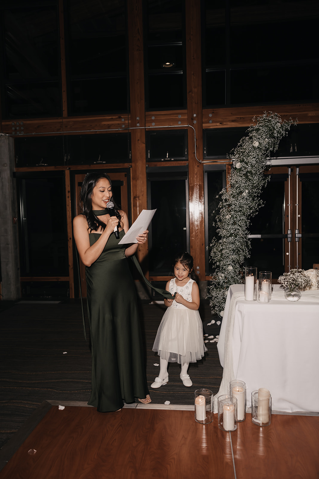 A maid of honor holding a microphone and reading a heartfelt speech, with a young flower girl standing beside her. Moments like these help create a meaningful flow in your wedding day timeline.