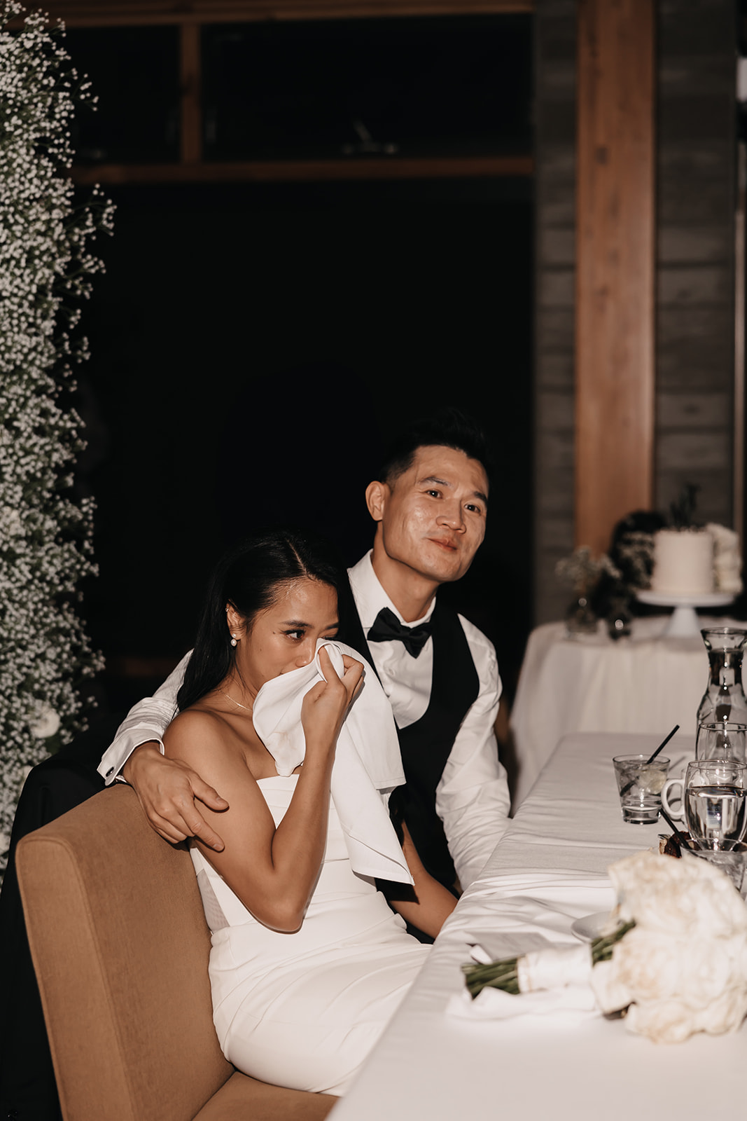 The couple shares an emotional moment during their wedding reception. The bride wipes away a tear while her groom lovingly supports her, capturing the tender essence of the Wedding Day Timeline.